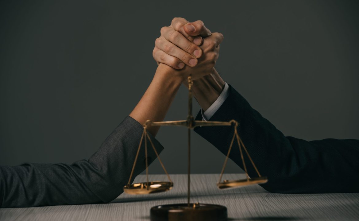 partial view of businessman and businesswoman arm wretsling on wooden table with scales of justice
