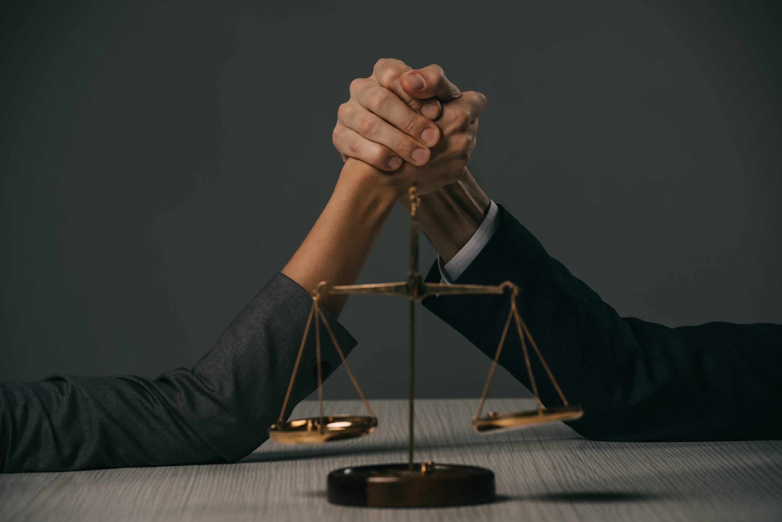 partial view of businessman and businesswoman arm wretsling on wooden table with scales of justice