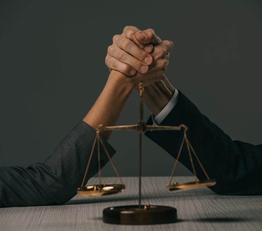 partial view of businessman and businesswoman arm wretsling on wooden table with scales of justice
