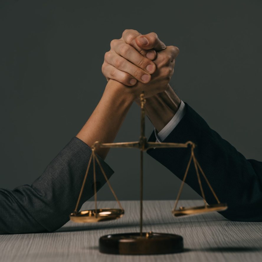 partial view of businessman and businesswoman arm wretsling on wooden table with scales of justice